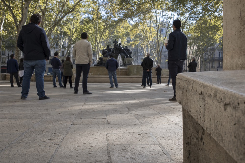 People stand scattered on a paved Paris square facing a bronze fountain under plane trees, with limestone buildings behind.