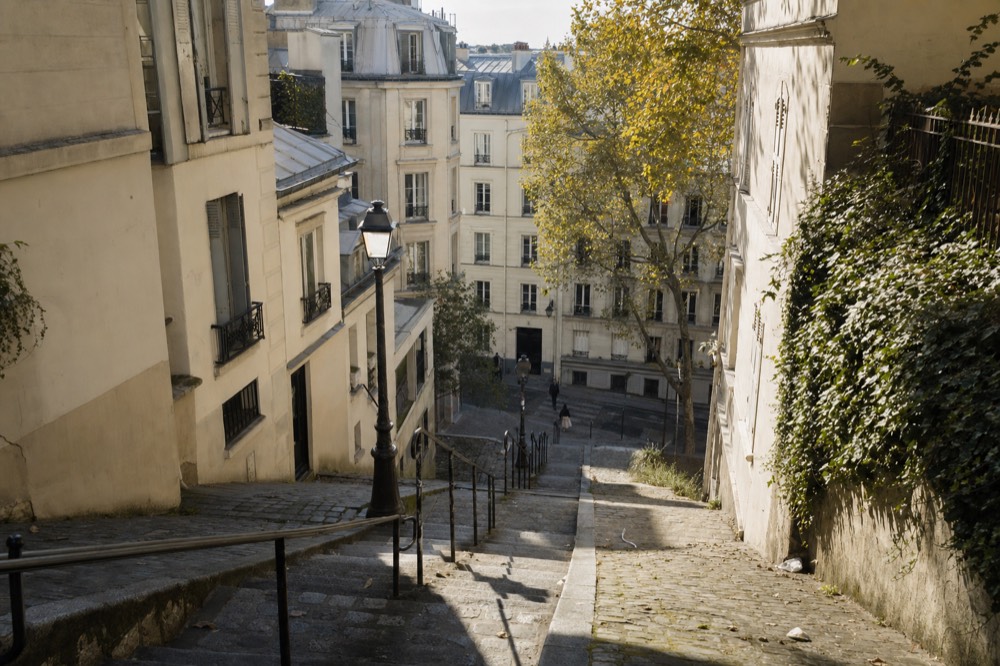 Steep cobbled Montmartre steps descend between cream buildings, iron rails, a lamp post, plane trees and an ivy-covered wall.