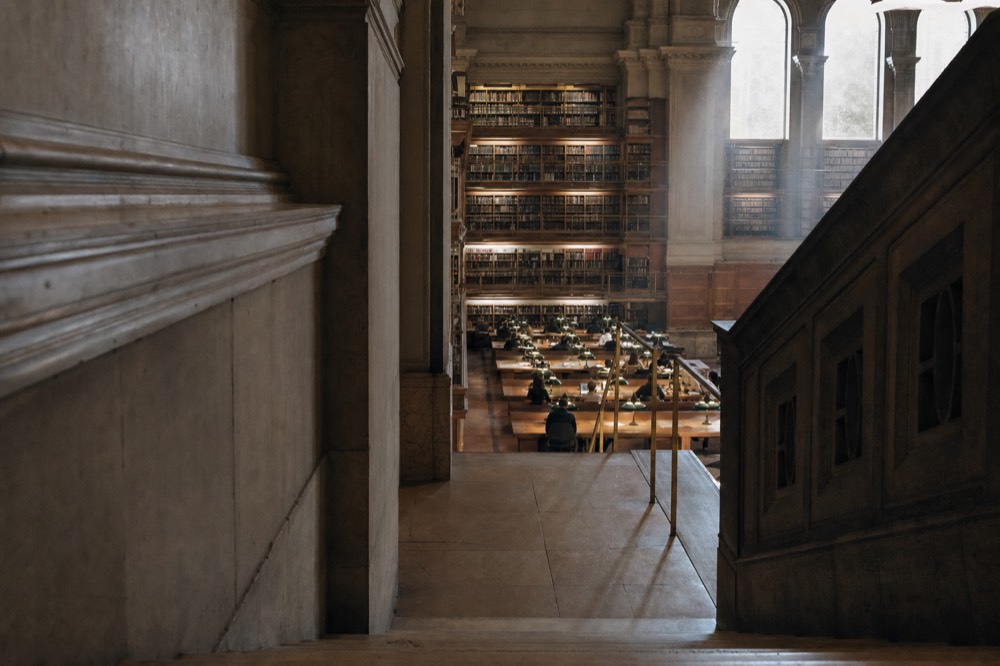 A grand library reading room seen from a stone stair, with tall bookshelves, long lamp-lit tables, arched windows, and distant readers.