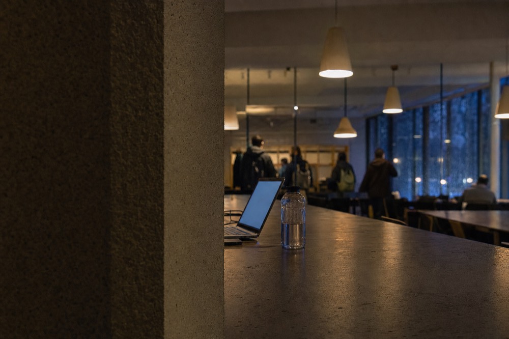 Warm pendant lights hang over long library tables with a laptop, water bottle, distant people, and rainy windows beyond.