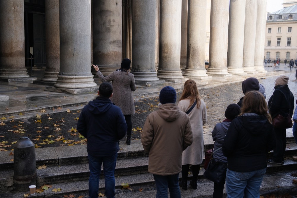 A guide addresses a small group from behind on wet stone steps beneath tall limestone columns with autumn leaves scattered nearby.