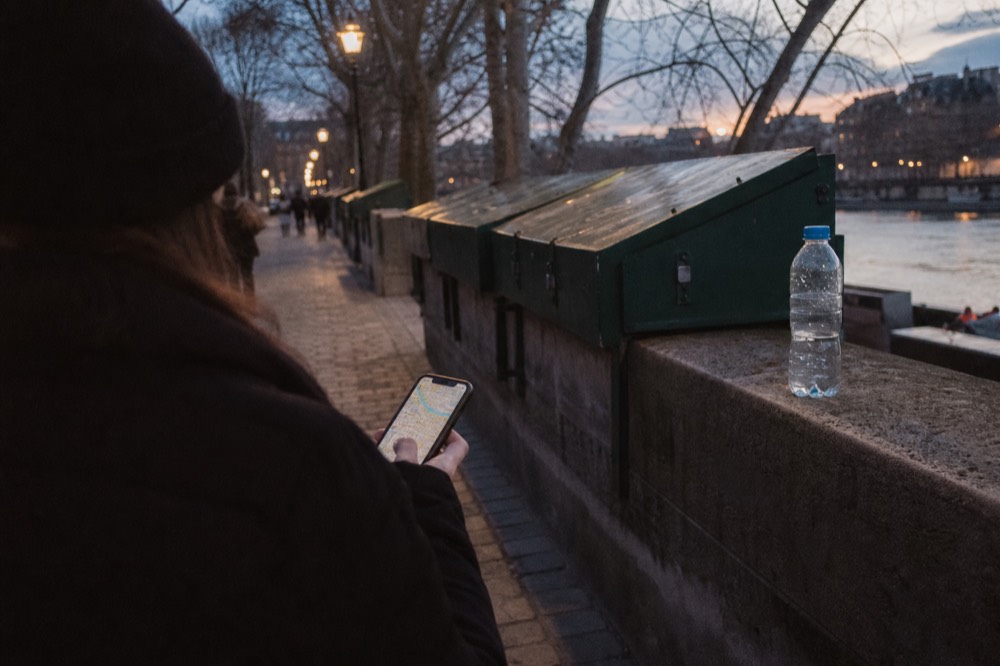 A walker looks at a lit phone map beside green bouquiniste boxes and a water bottle on a Seine quay at dusk.