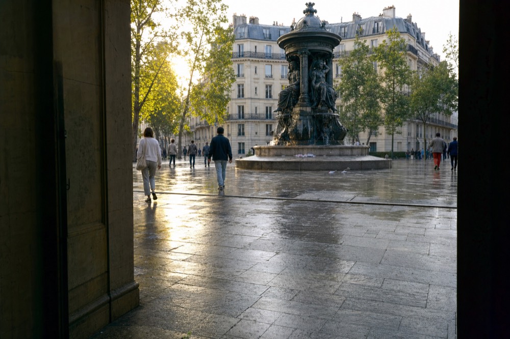 People walk across wet paving around a dark bronze fountain in a Paris square framed by a shadowed doorway.