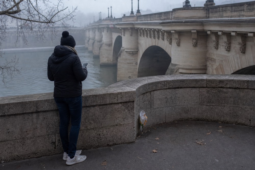 A lone walker checks a phone by a stone parapet beside the misty Seine, with pale arched bridge spans and a wrapper nearby.