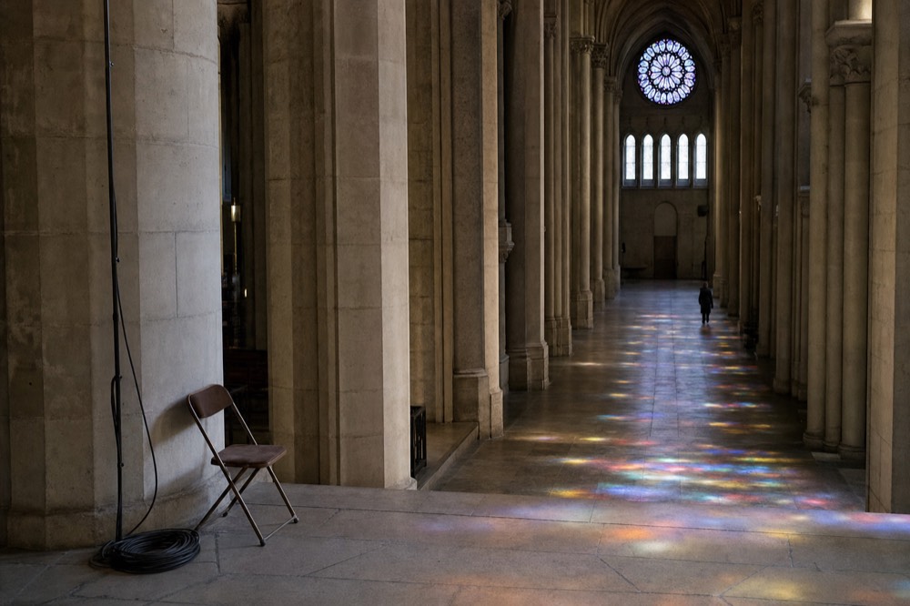 Gothic cathedral aisle with tall stone columns, a rose window, colored light on the floor, a folding chair, and a coiled cable.