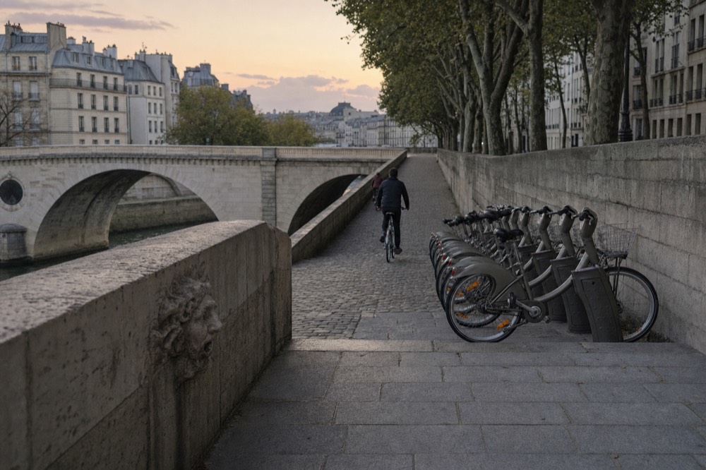 Docked city bikes line a cobbled Seine quay beside a stone bridge, with a cyclist riding away under plane trees at dusk.