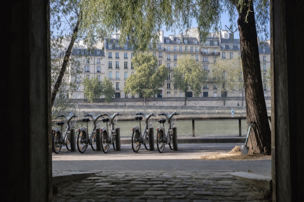 City bicycles stand in a docking rank beside a tree-lined Paris riverbank with cream stone apartments across the water.