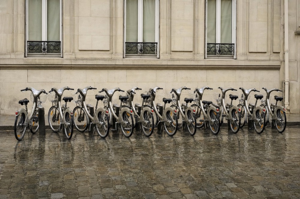 City bicycles stand in a row on wet cobblestones against a pale stone Paris facade.