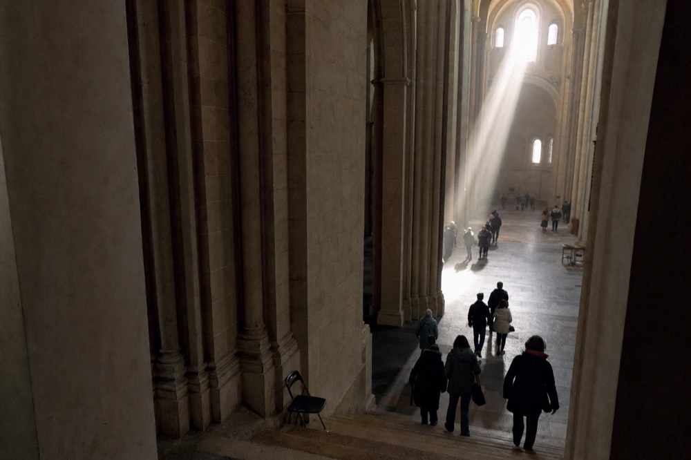 Sunlight streams through high church windows onto stone arches and a floor where distant visitors walk down steps.
