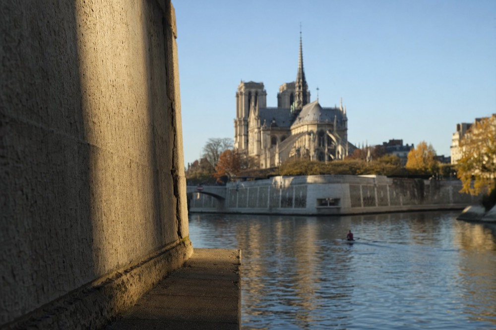Notre-Dame rises beyond the Seine at dusk, seen past a close stone wall with its spire, buttresses, river and autumn trees visible.