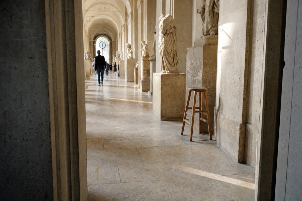 A long arched museum gallery with stone statues on plinths, a polished floor, distant visitors, a clock window, and a wooden stool.