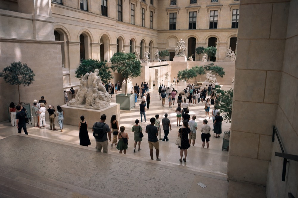 Visitors stand on steps and gather around marble sculptures in a large stone museum courtyard with arched walls and indoor trees.