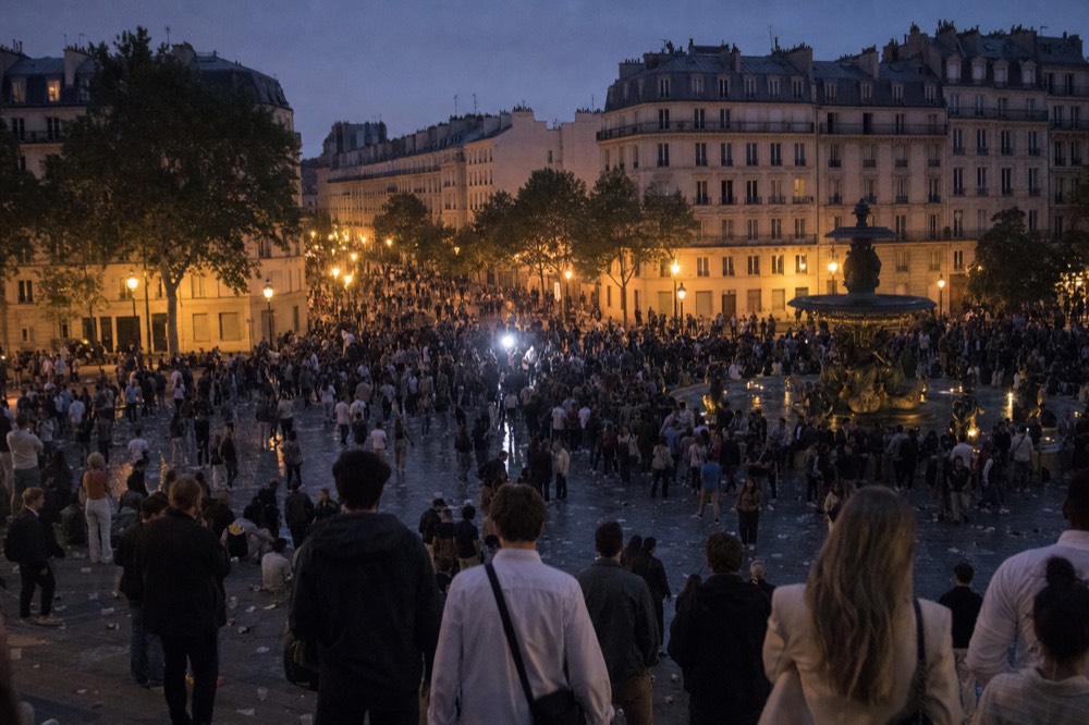 A dense evening crowd fills a Paris square around a fountain, with limestone buildings and trees under a violet dusk sky.