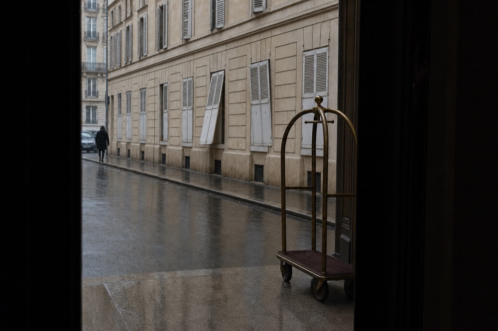 Rainy Paris street seen from a dark doorway, with cream shuttered facades, a distant pedestrian, and a porter trolley by the entrance.