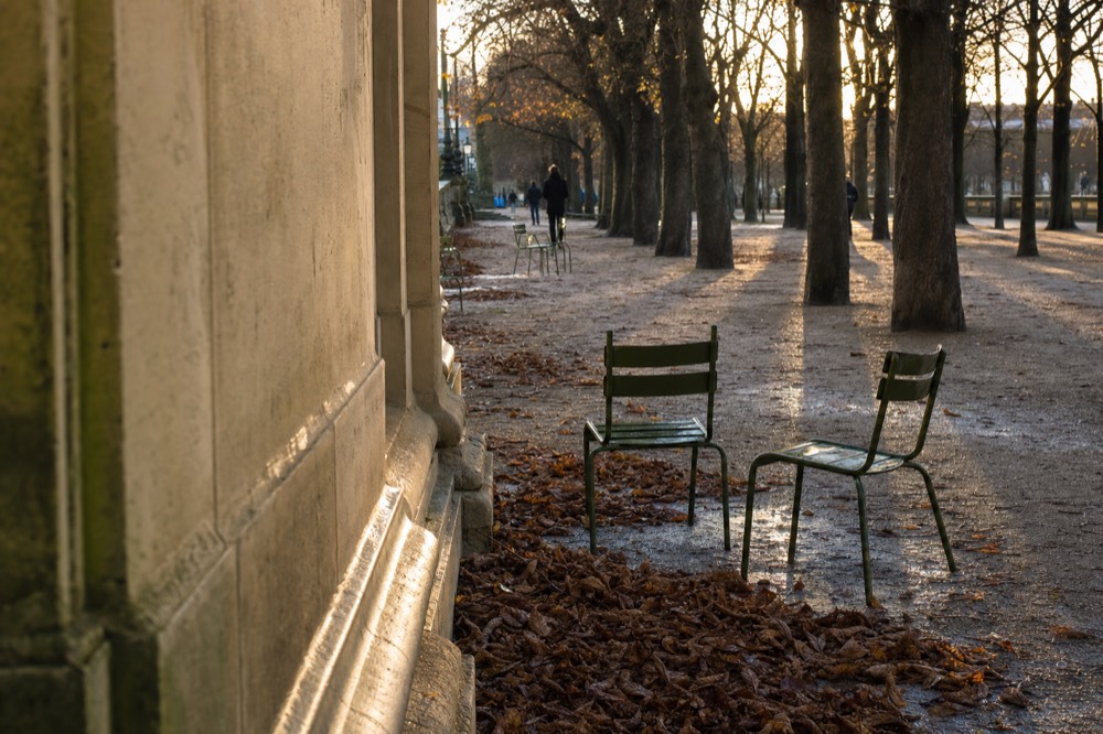 Green metal chairs stand beside heaps of fallen leaves on a wet gravel path under bare trees by a stone wall.