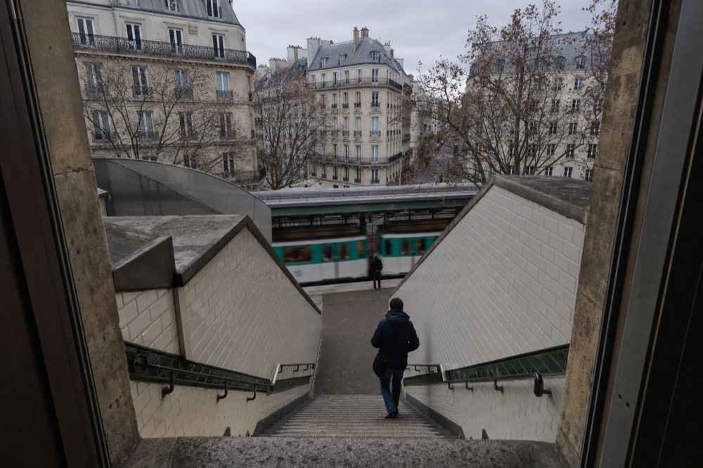 A tiled metro stairway descends toward an elevated platform where a green and white train passes, with Paris apartment blocks beyond.