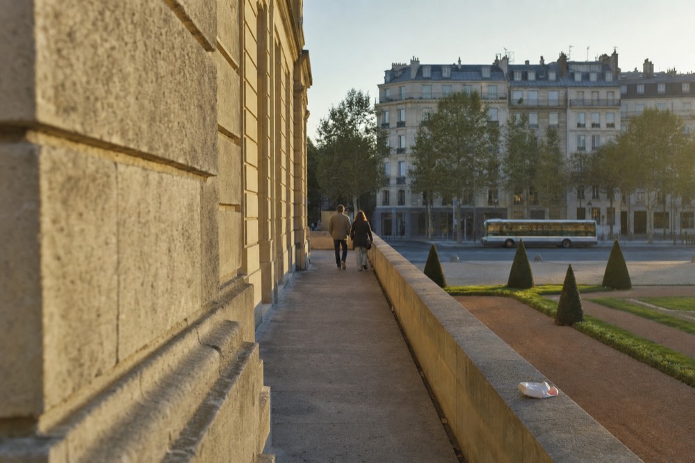 A city bus passes a broad Paris boulevard beyond a sunlit stone terrace, trimmed garden beds, distant pedestrians, and Haussmann buildings.
