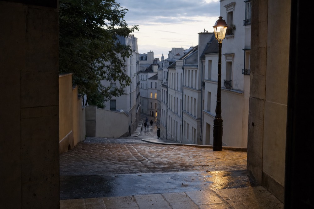 Wet cobbled Paris street slopes downhill between pale buildings, with a lit cast-iron lamp and distant pedestrians at dusk.