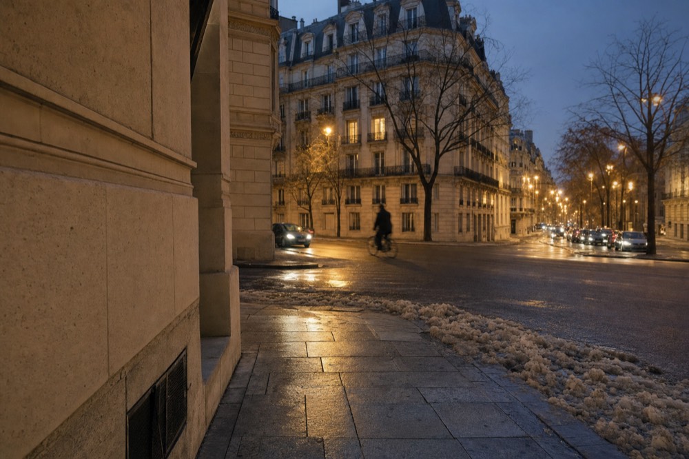 Wet Paris boulevard at dusk with Haussmann buildings, streetlights, bare trees, slush along the curb and a blurred cyclist crossing.