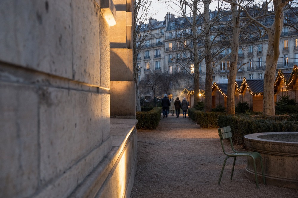 Dusk view of lit wooden Christmas market chalets beside hedges, plane trees, a stone basin and a green chair in Paris.
