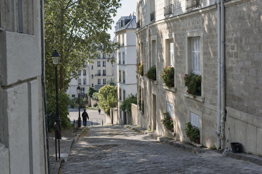 A cobbled sloping Paris street lined with limestone buildings, shuttered windows, geranium planters, lampposts, trees and two distant pedestrians.