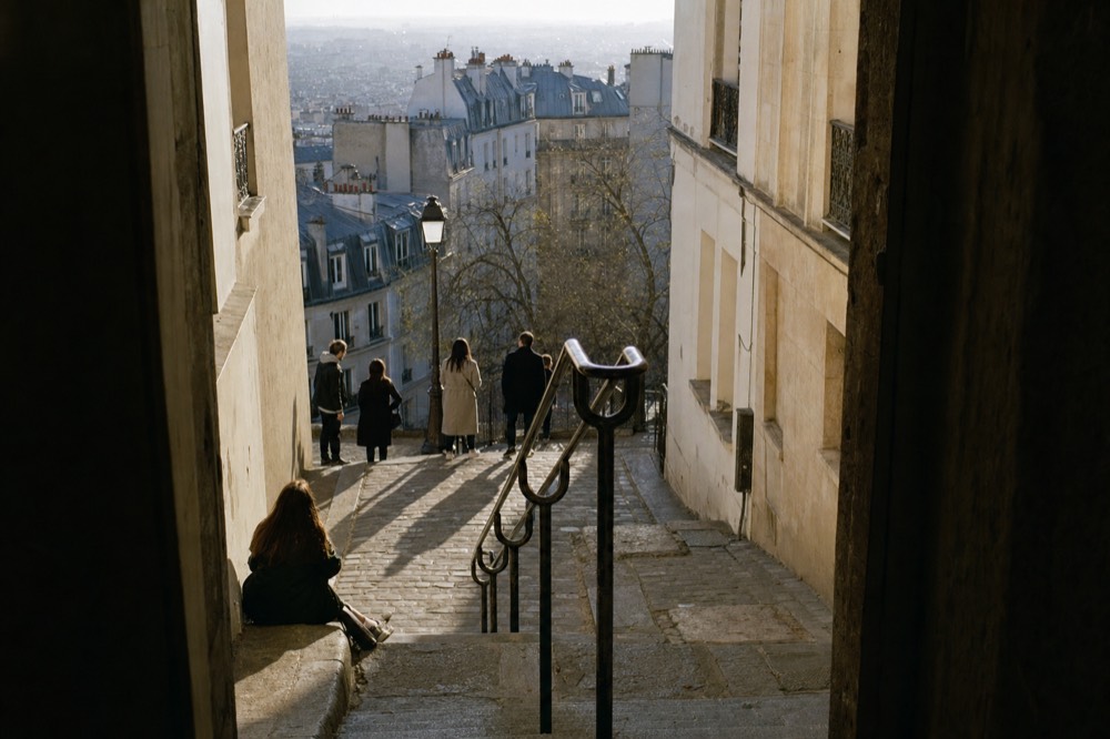 People pause on a steep stone stairway between pale Paris buildings, with grey mansard rooftops and hazy city views below.