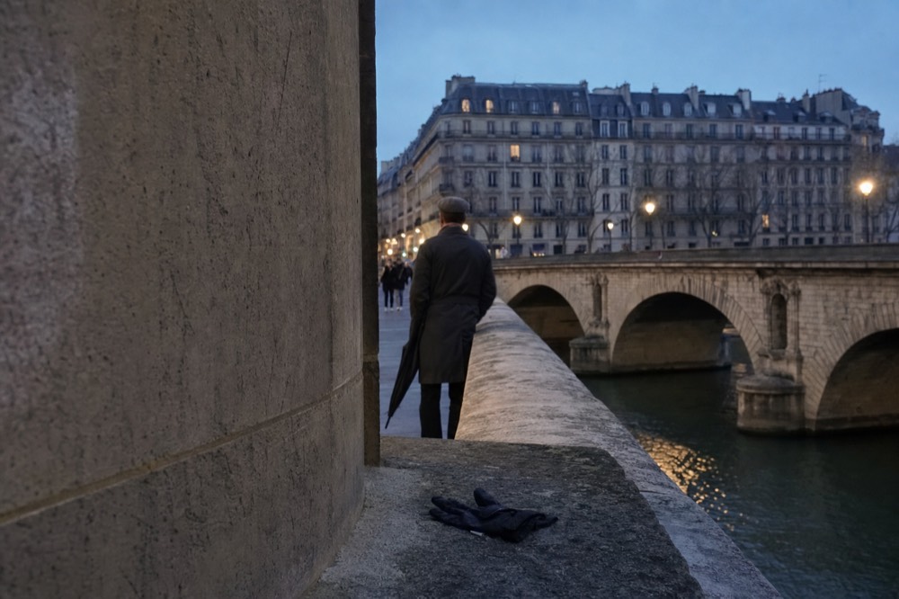 A person with a furled umbrella stands by a stone parapet on a Paris bridge at dusk, with a glove on the wall and river below.