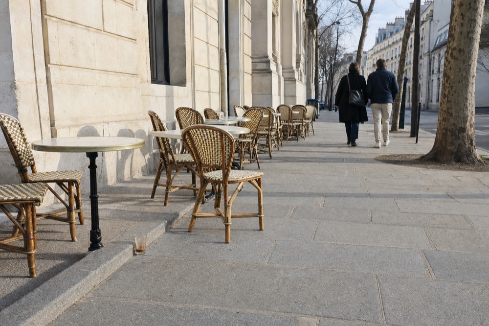 Rattan cafe chairs and small round tables line a wide Paris pavement beside a cream stone facade and plane trees.