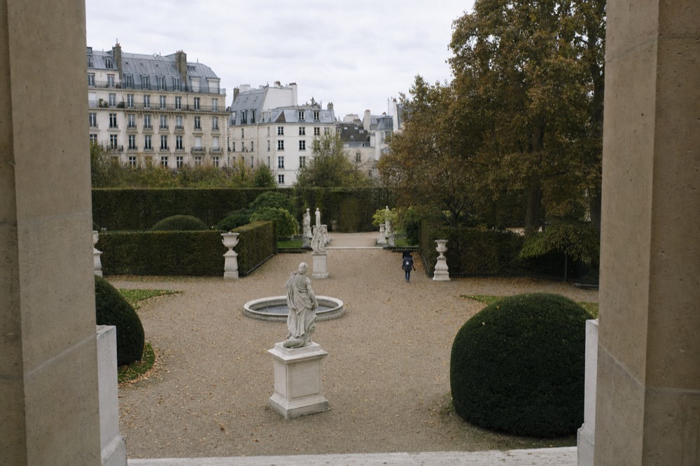 A stone arcade overlooks a Parisian gravel courtyard with classical statues, round basins, clipped hedges, trees, and mansard-roof buildings.