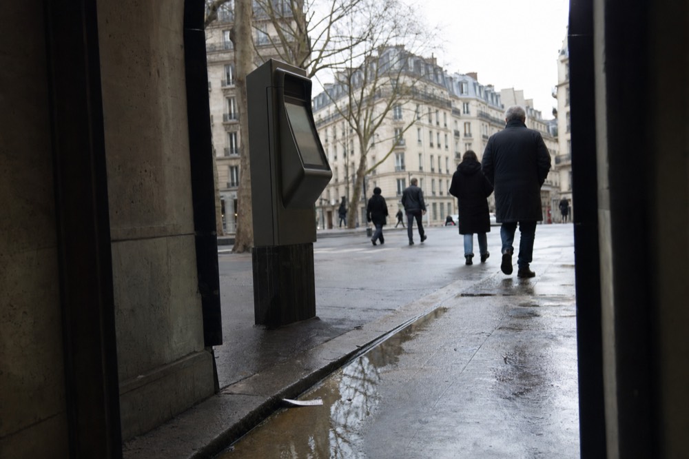 A blank ticket machine stands by a wet Paris pavement with pedestrians and Haussmann buildings beyond.