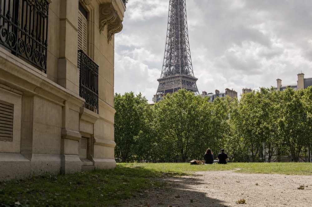 Two distant people sit on a lawn by a gravel path, with plane trees and the Eiffel Tower rising behind a Parisian building.