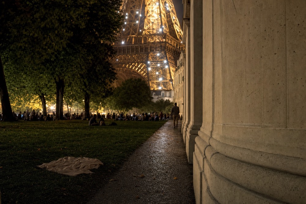 The Eiffel Tower sparkles at night beyond dark trees, a wet path, a stone wall, distant people, and a blanket on the grass.