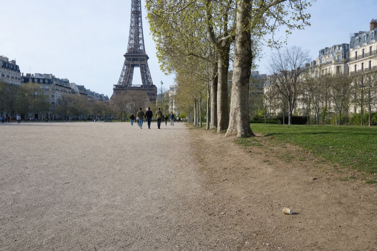 The Eiffel Tower rises beyond a wide gravel walk lined with plane trees, Paris buildings, distant pedestrians and a paper cup.