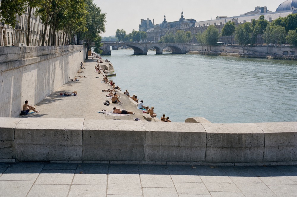 People lounge on a sunny stone quay beside the Seine, with a bridge, trees, and grand Parisian buildings in the distance.