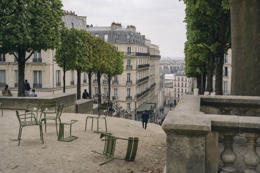 Green metal chairs sit on a gravel terrace under clipped trees, overlooking cream Paris buildings and a descending street.