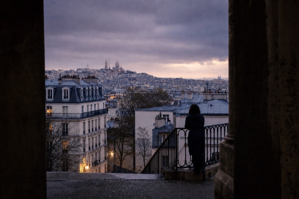 Dusk view over Paris rooftops toward Sacre-Coeur, with a silhouetted person leaning by a wrought-iron terrace railing.