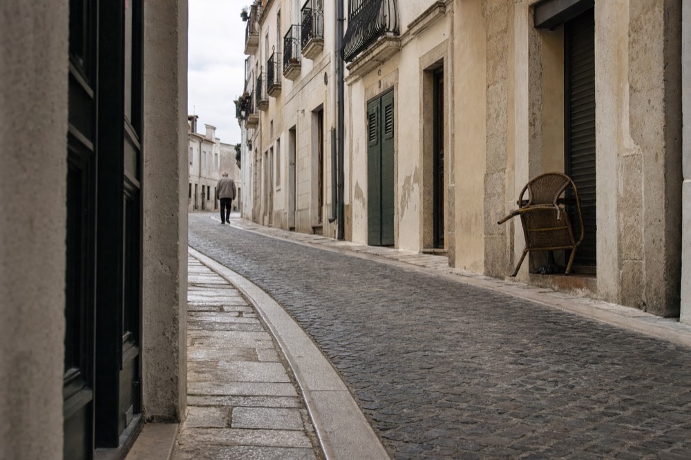 Narrow cobbled street with cream limestone fronts, shuttered doors, wrought-iron balconies, a stacked chair, and one distant walker.