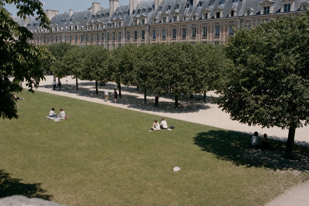 People sit on a sunny Paris park lawn below clipped trees and brick arcaded buildings with grey mansard roofs.