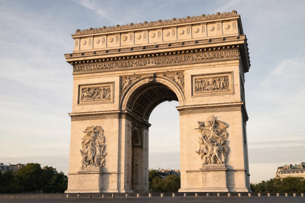 The Arc de Triomphe stands in warm light with trees and distant rooftops behind it.