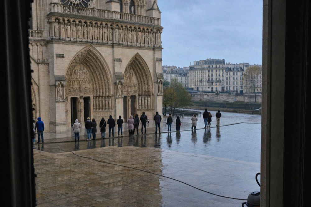 Visitors queue on the wet parvis before Notre-Dame cathedral, seen from a dark opening with Paris buildings beyond the Seine.