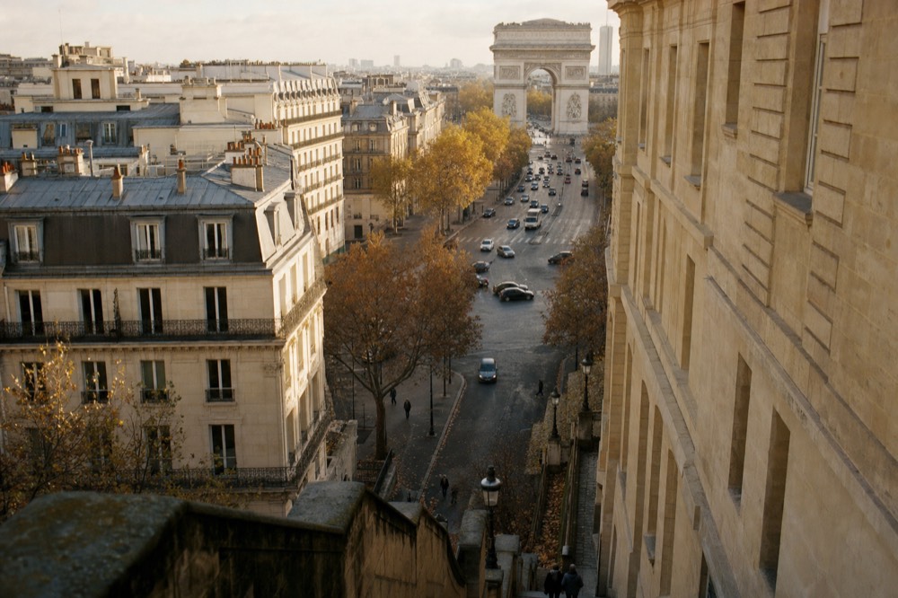 Elevated view over Haussmann buildings and autumn trees toward the Arc de Triomphe with traffic on a Paris avenue.