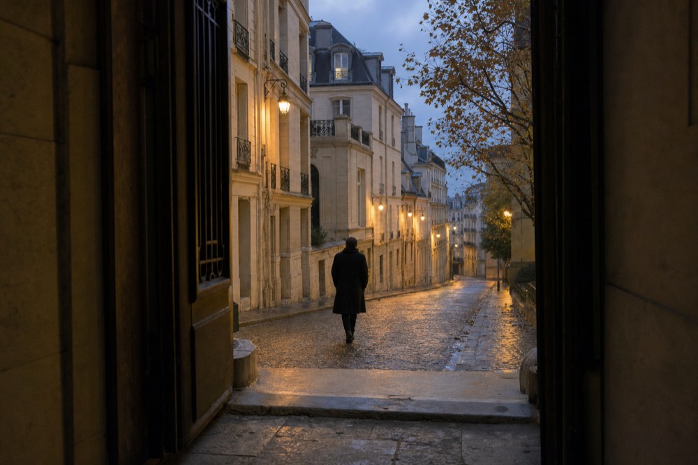 A wet cobbled Paris lane at dusk is seen from a dark doorway, with lamplit stone buildings and a lone figure walking away.