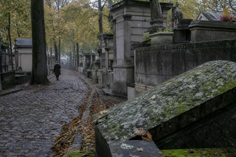 A tree-shaded cobbled avenue of weathered tombs and mossy stone at Pere Lachaise cemetery on a misty November morning, free to walk into.