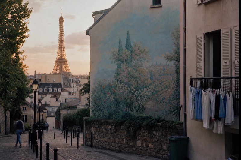 The Eiffel Tower catching warm dusk light above Paris zinc rooftops, seen for free down a stepped side street past a painted mural on a gable wall.