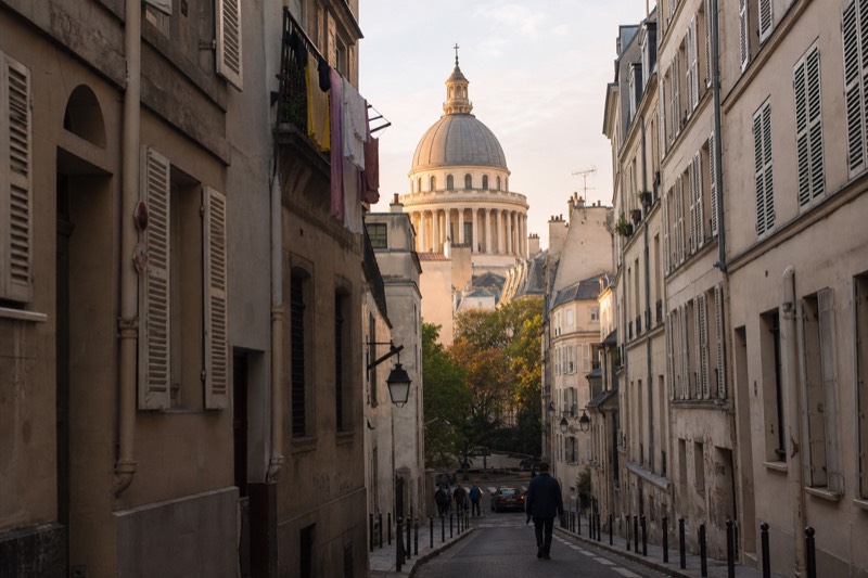 A narrow Latin Quarter lane in the 5th arrondissement at golden hour, the dome of the Pantheon rising above the rooftops at the end of the street.