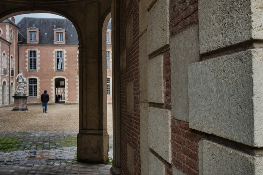 Arcaded stone walkway opens onto a gravel courtyard with rose brick walls, pale stone quoins, a statue, cobbles, puddles, and distant visitors.
