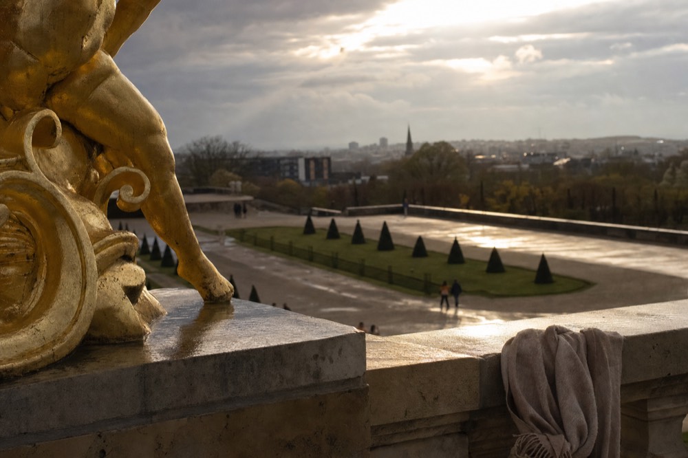 A gilded statue and scarf on a stone terrace overlook wet garden paths and a distant Paris skyline under storm clouds.