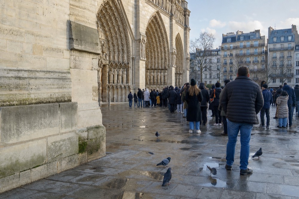 People queue beside Notre-Dame's carved limestone portals on a wet parvis with puddles and pigeons in morning light.