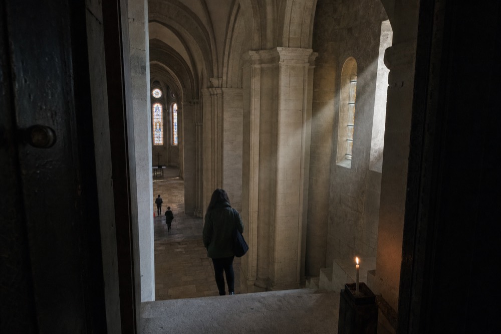 Stone church nave seen from a doorway with tall columns, stained glass windows, a candle, sunbeams, and distant visitors.
