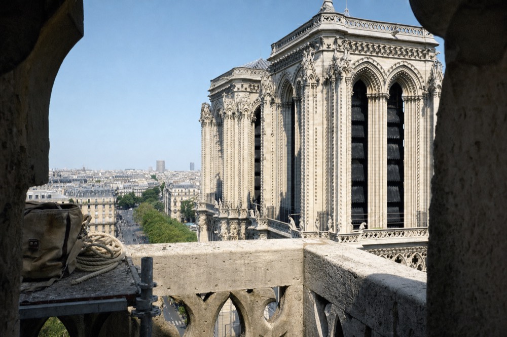 Gothic bell towers rise above a Paris skyline, seen from a shadowed stone opening with a tool bag and coiled rope on a ledge.